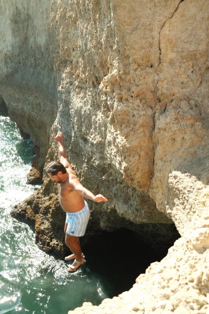 A man jumping into the turquoise waters below the Algarve cliffs, surrounded by dramatic coastal rock formations in southern Portugal.