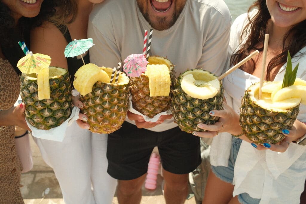 Friends holding tropical cocktails served inside fresh pineapples during a sunny day in Portugal.