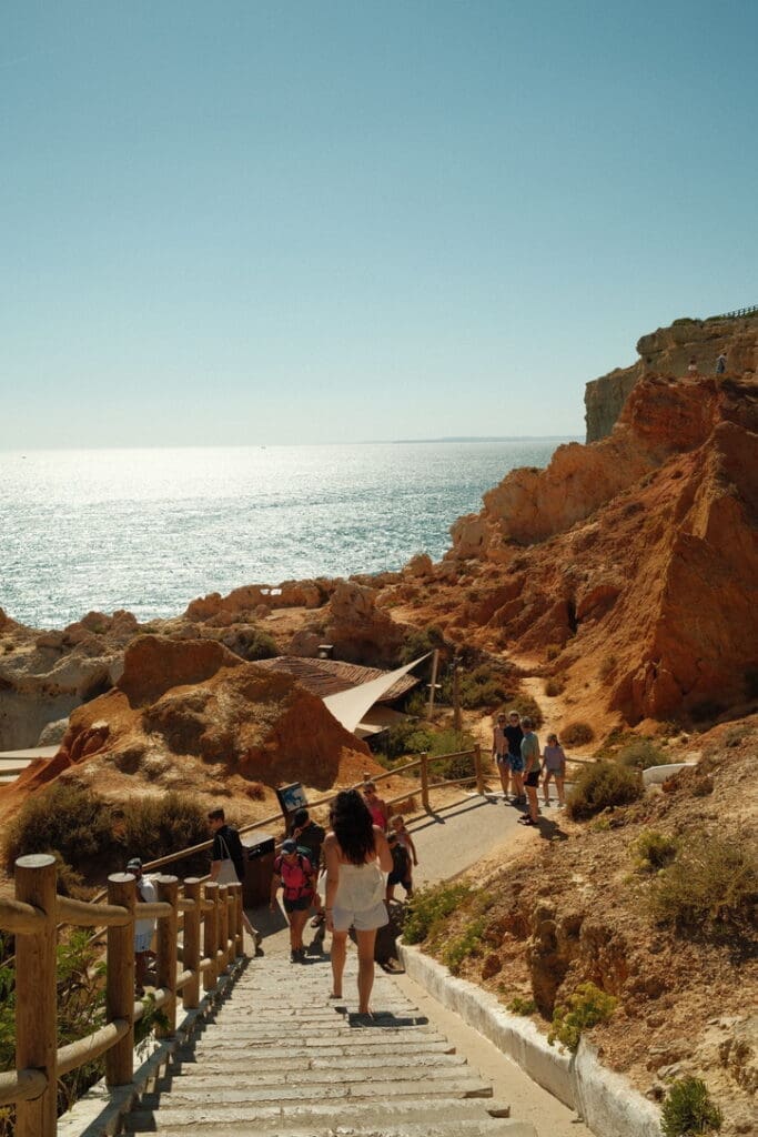 Visitors walking down a staircase toward the rugged Algarve coastline, surrounded by golden cliffs and panoramic ocean views.