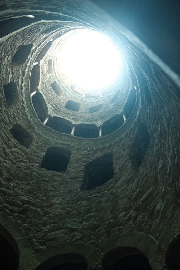 Looking up through the circular stone interior of the historic Quinta da Regaleira estate toward the sky above.
