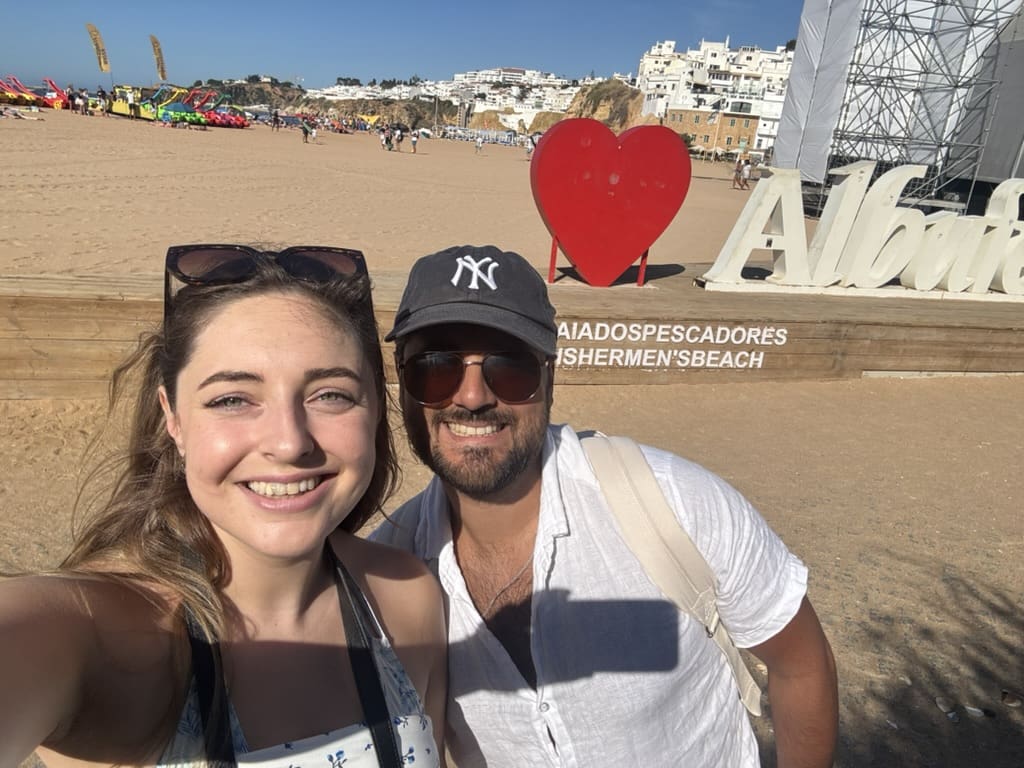 A couple smiling at Fisherman's Beach in Albufeira, Portugal, with the beach, cliffs, and iconic "I ❤️ Albufeira" sign in the background.