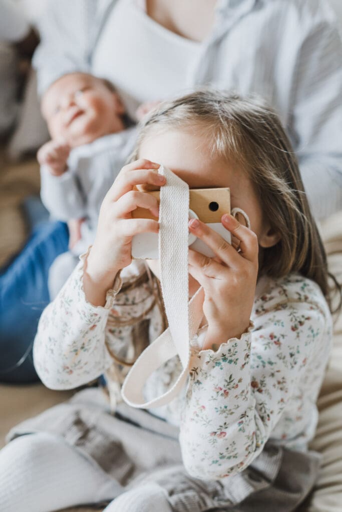 Older sister holding a toy camera and pretending to take photos during a playful at-home newborn and family session.