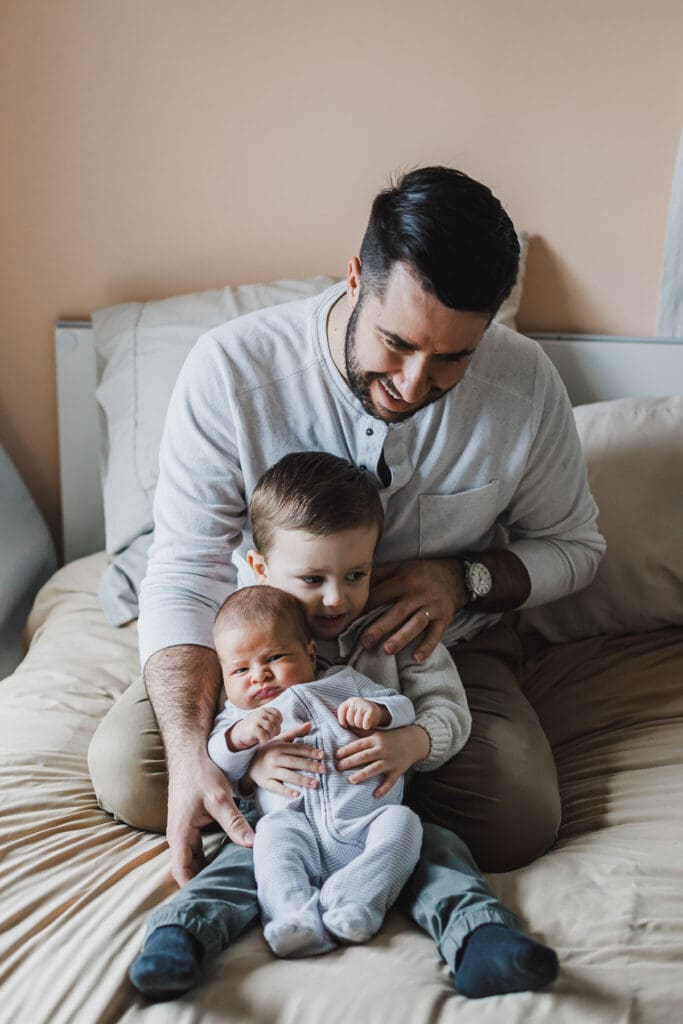 Father sitting on a bed with his newborn baby while the older siblings gather close during a relaxed family moment at home.