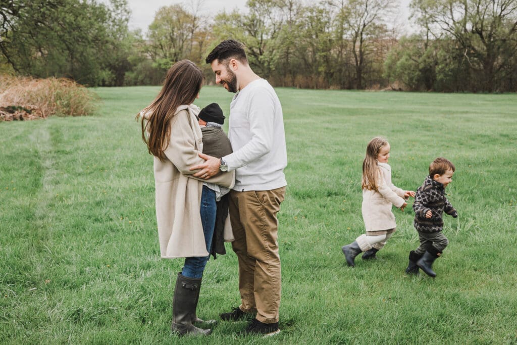 Parents sharing a quiet moment with their newborn wrapped in a carrier while their two young children run and play together in a grassy backyard during a cozy spring family session.
