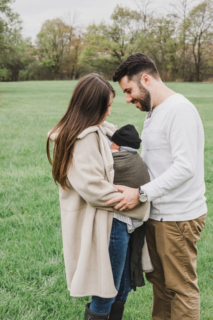 Parents smiling at their newborn baby while standing together in a grassy field during an outdoor family session.