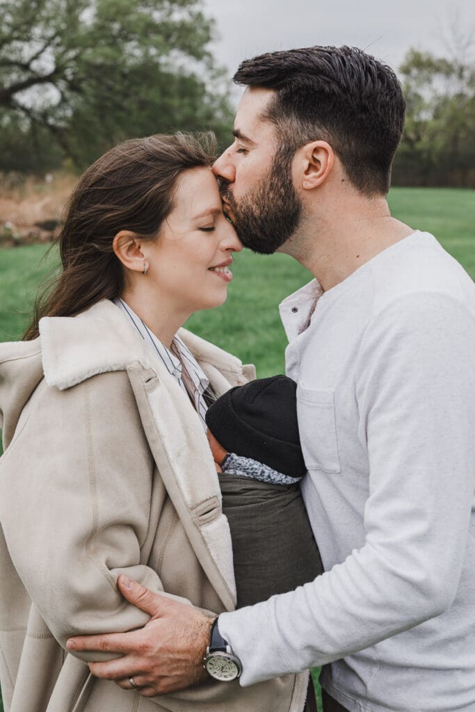 Father kissing mother on the forehead while they hold their newborn baby during a cozy outdoor newborn and family session.