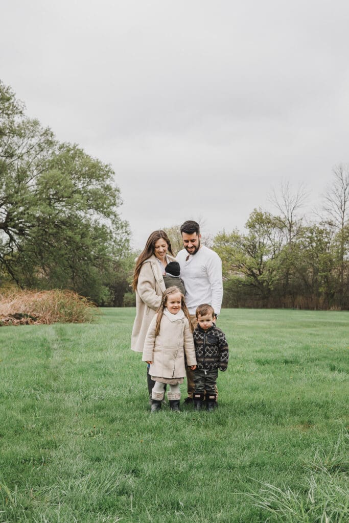 The Turner family standing together in a grassy field with their newborn baby and two young children during an outdoor family session.
