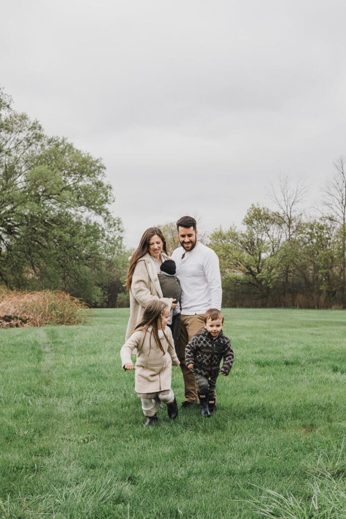 Parents holding their newborn while their two children run and play in front of them during a relaxed outdoor family session.
