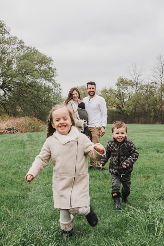 Two young siblings running toward the camera while their parents and newborn baby smile in the background during a family session.