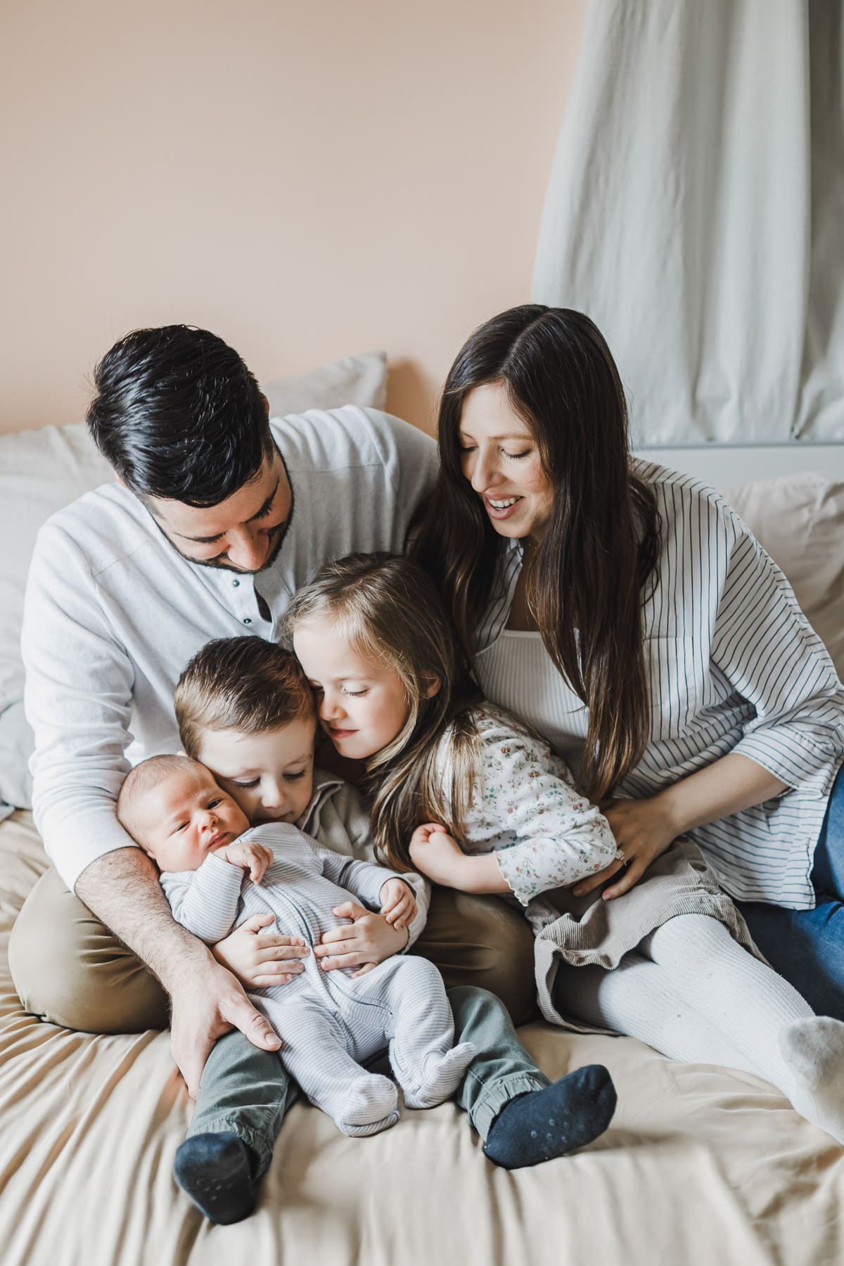 Parents and their three children cuddled together on a bed at home, with the older siblings gently holding and admiring their newborn baby during a cozy lifestyle newborn and family session.