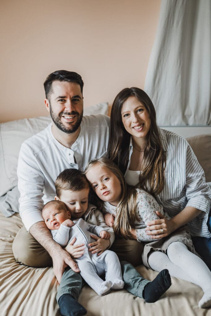 Parents and their three children cuddled together on a bed at home, with the older siblings gently holding and admiring their newborn baby during a cozy lifestyle newborn and family session.