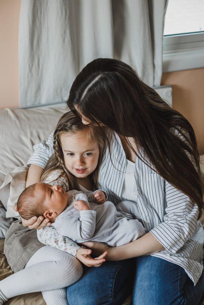 Mother cuddling her newborn baby while the older sister looks on during a relaxed family session at home.