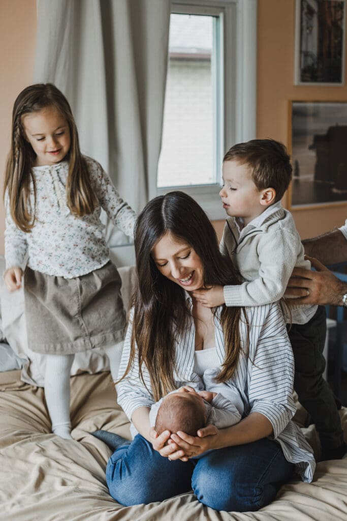 Older siblings standing beside their mother and newborn baby, sharing a sweet family moment during an at-home newborn session.